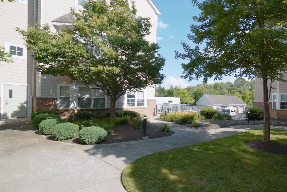 Outdoor view of a senior living facility showing a paved walkway surrounded by green grass, bushes, and trees. The building exterior is beige with white trim and multiple windows. The sky is clear with some clouds.