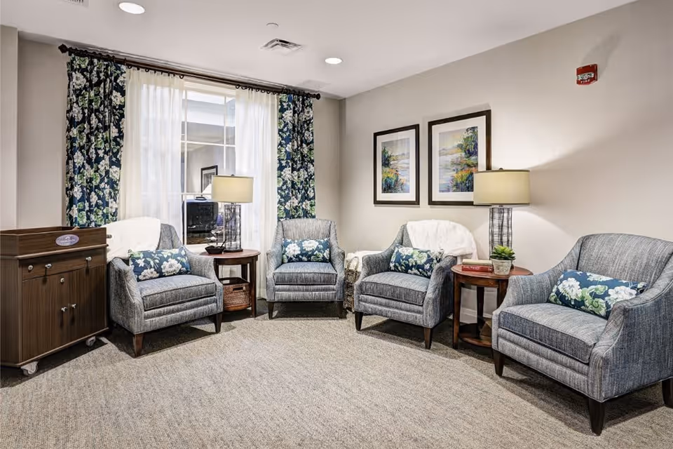 A cozy seating area with four gray upholstered armchairs, each with a floral cushion. Two wooden side tables with lamps and decorative items are placed between the chairs. Behind the chairs is a window with floral curtains and sheer white drapes. Two framed landscape paintings hang on the beige wall.