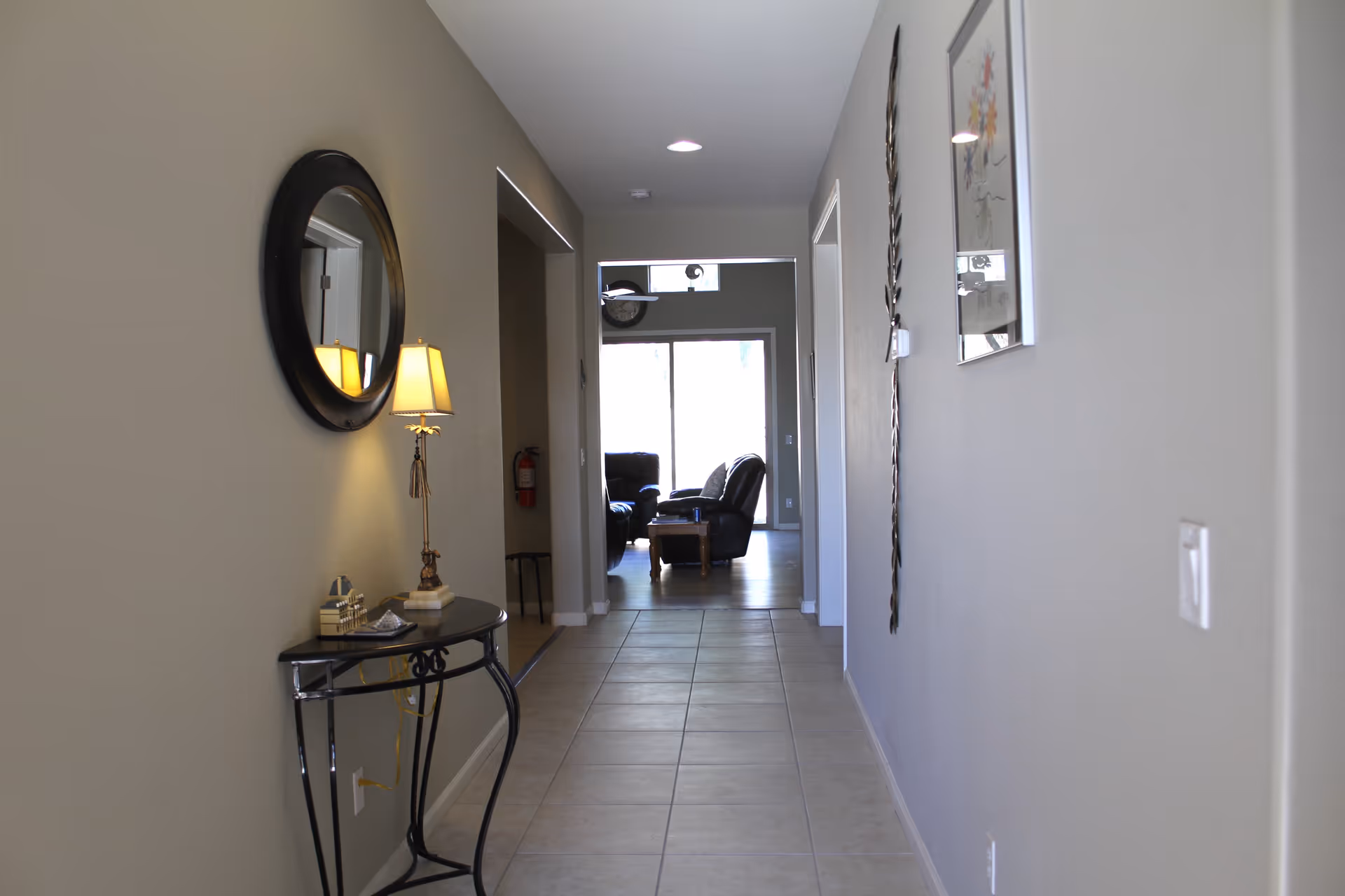 A hallway with beige tiled floor and light-colored walls. On the left side, there is a small black table with decorative items and a lamp, and a round mirror above it. On the right wall, there is a framed picture and a decorative wall piece. At the end of the hallway, a living room area is visible with two black leather chairs and a wooden coffee table in front of a large window or sliding glass door.