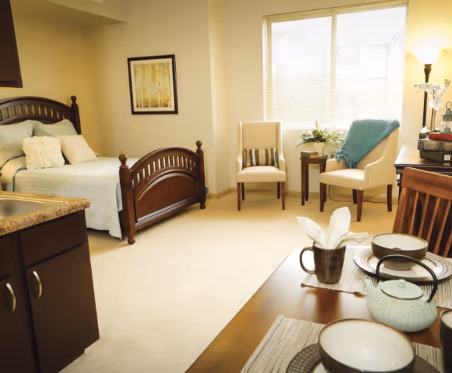 A cozy and well-lit room featuring a wooden bed with light blue bedding and pillows, two beige chairs with a small table and plant between them near a window with blinds, a wooden dining table set with bowls, cups, and a teapot, and a kitchen counter with cabinets in the foreground.