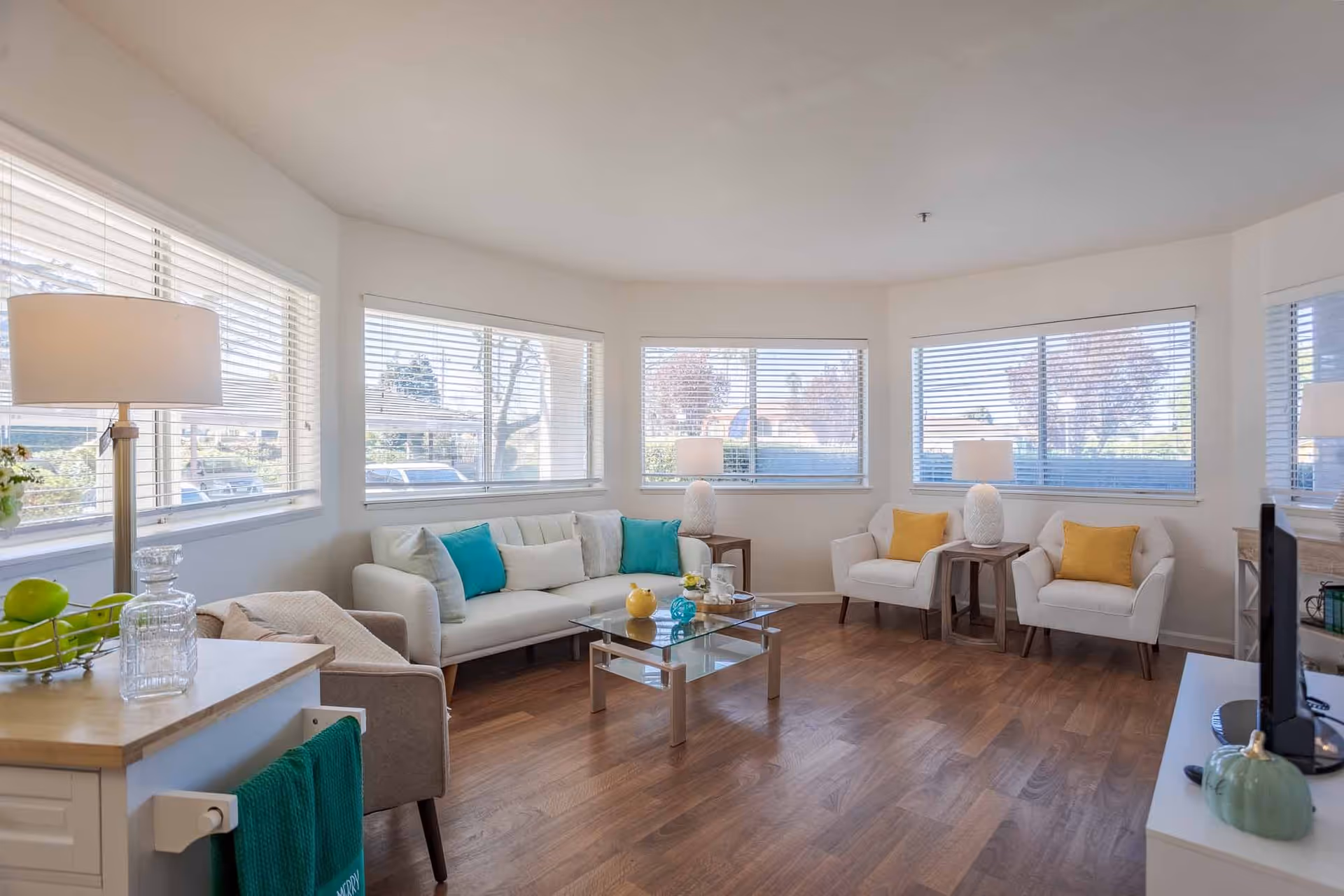 A bright and spacious living room with large windows on three sides letting in natural light. The room features a white sofa with teal and white cushions, two white armchairs with yellow cushions, a glass coffee table with decorative items, wooden flooring, and several table lamps. A small kitchen counter with a lamp, green apples, and a green towel is visible in the foreground.