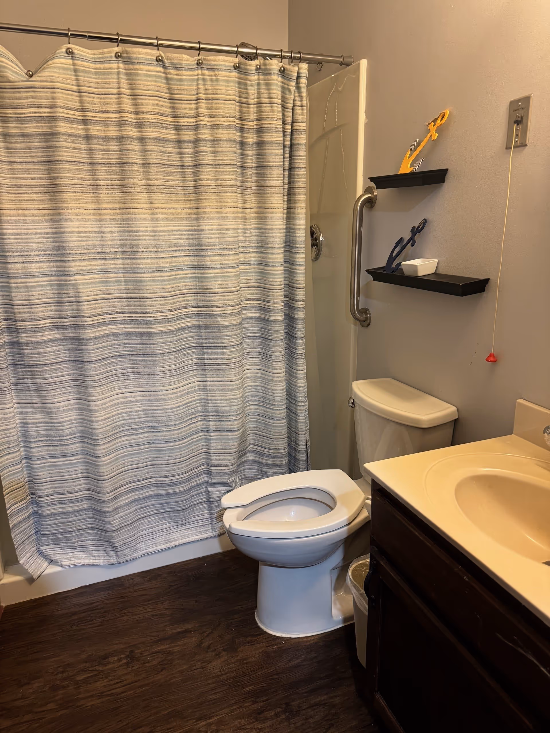 A bathroom with a white toilet, a sink with a beige countertop and dark wooden cabinet, a shower with a striped blue and white curtain, and two small black shelves on the wall holding decorative anchors and a small white dish. There is a grab bar next to the shower and a pull cord hanging from the wall.