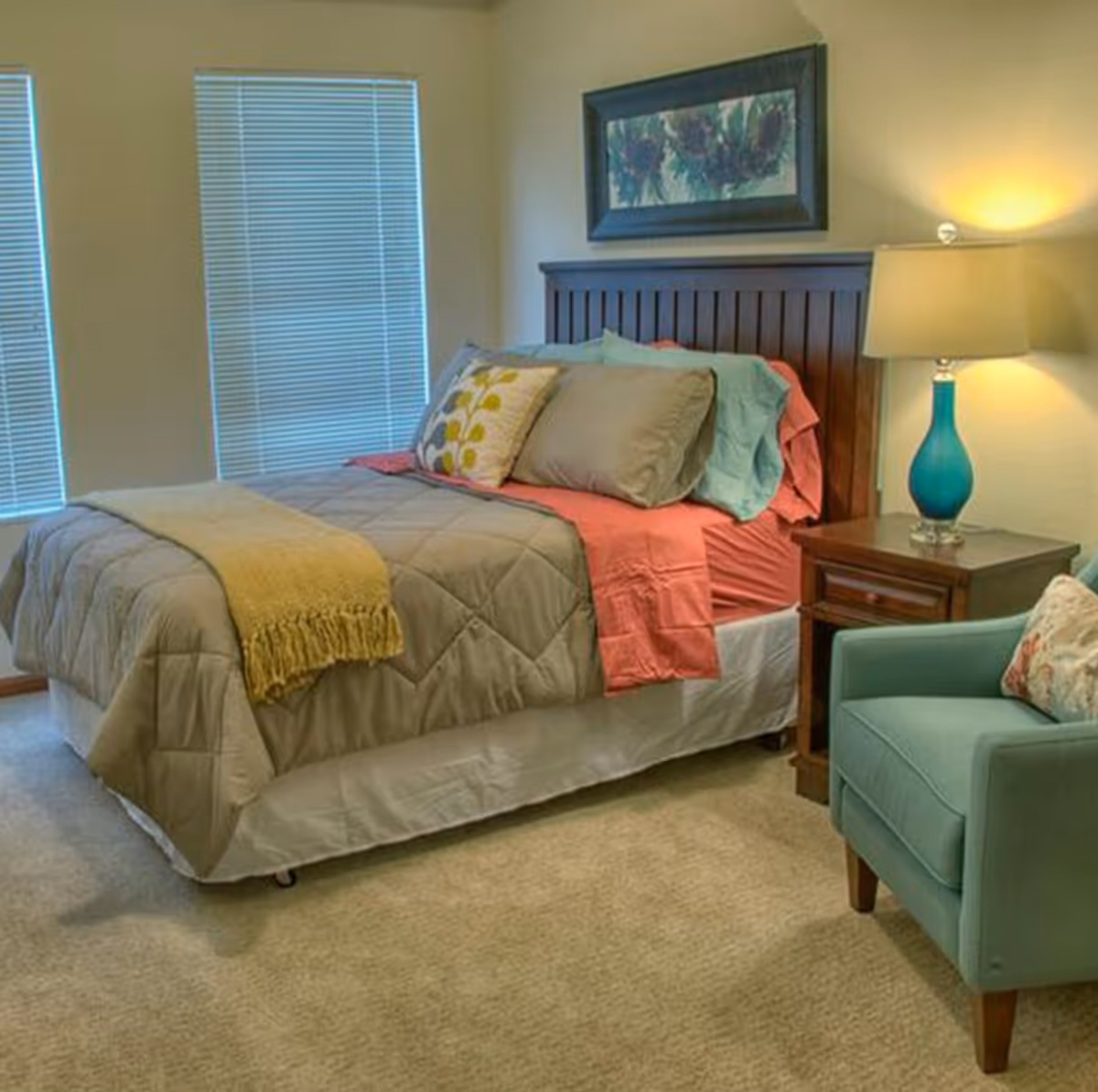 A cozy bedroom with a neatly made bed featuring a gray comforter, coral sheets, and multiple pillows. There is a wooden headboard, a nightstand with a blue lamp, and a teal armchair with a floral pillow. Two windows with closed blinds are visible on the left wall, and a framed floral painting hangs above the bed.