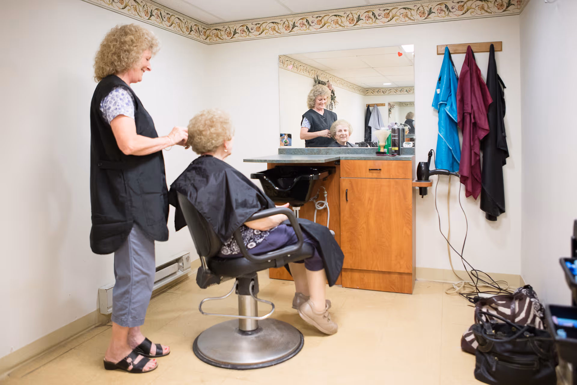 A hairdresser styling an elderly woman's hair in a salon area with a large mirror, wooden cabinet, and hairdressing tools. Several hairdressing capes hang on hooks on the wall.