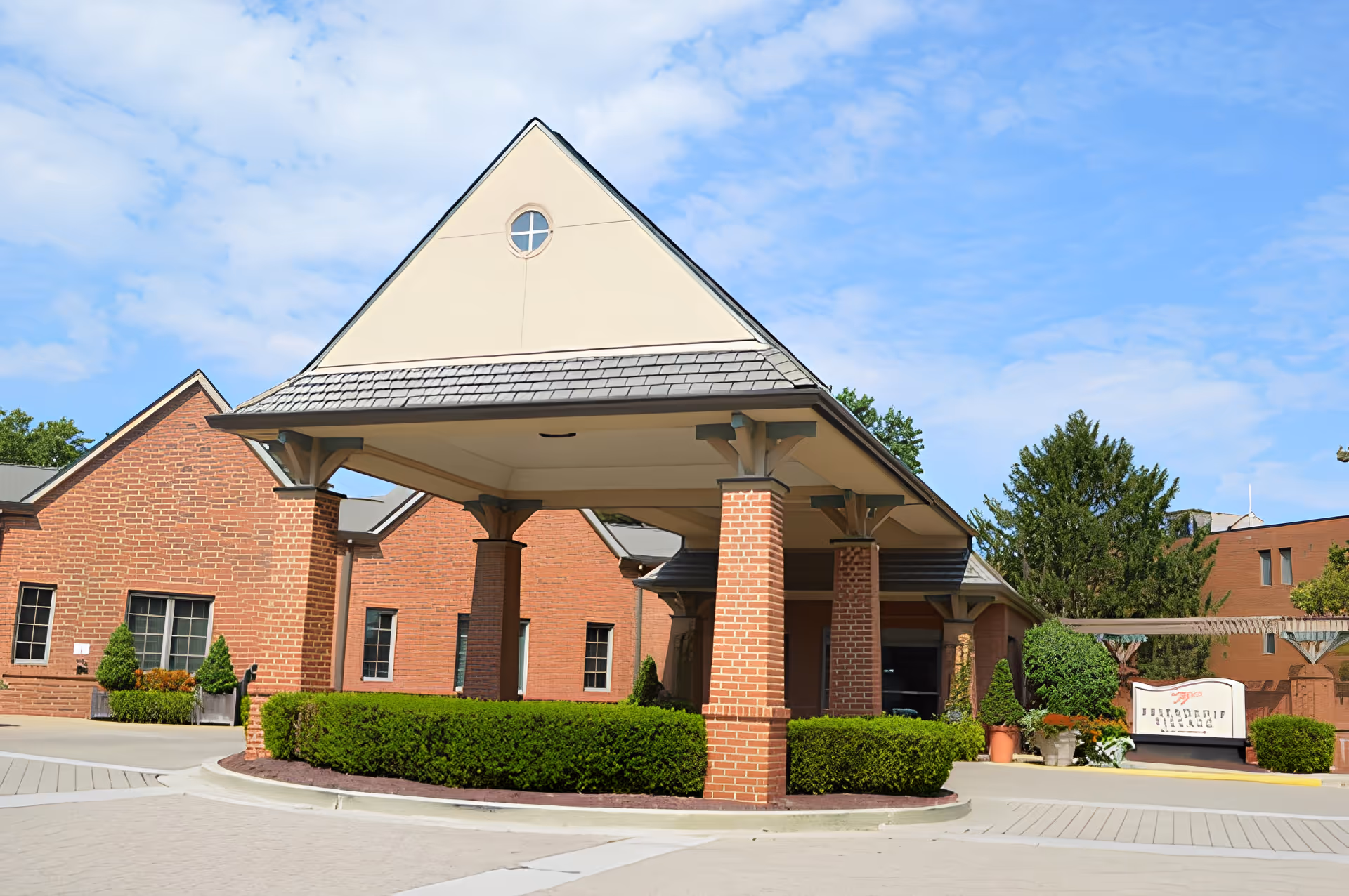 Exterior view of Friendship Village Chesterfield showing a covered entrance with brick pillars and a triangular roof. The building is made of red brick with several windows, surrounded by green bushes and trees under a partly cloudy blue sky.