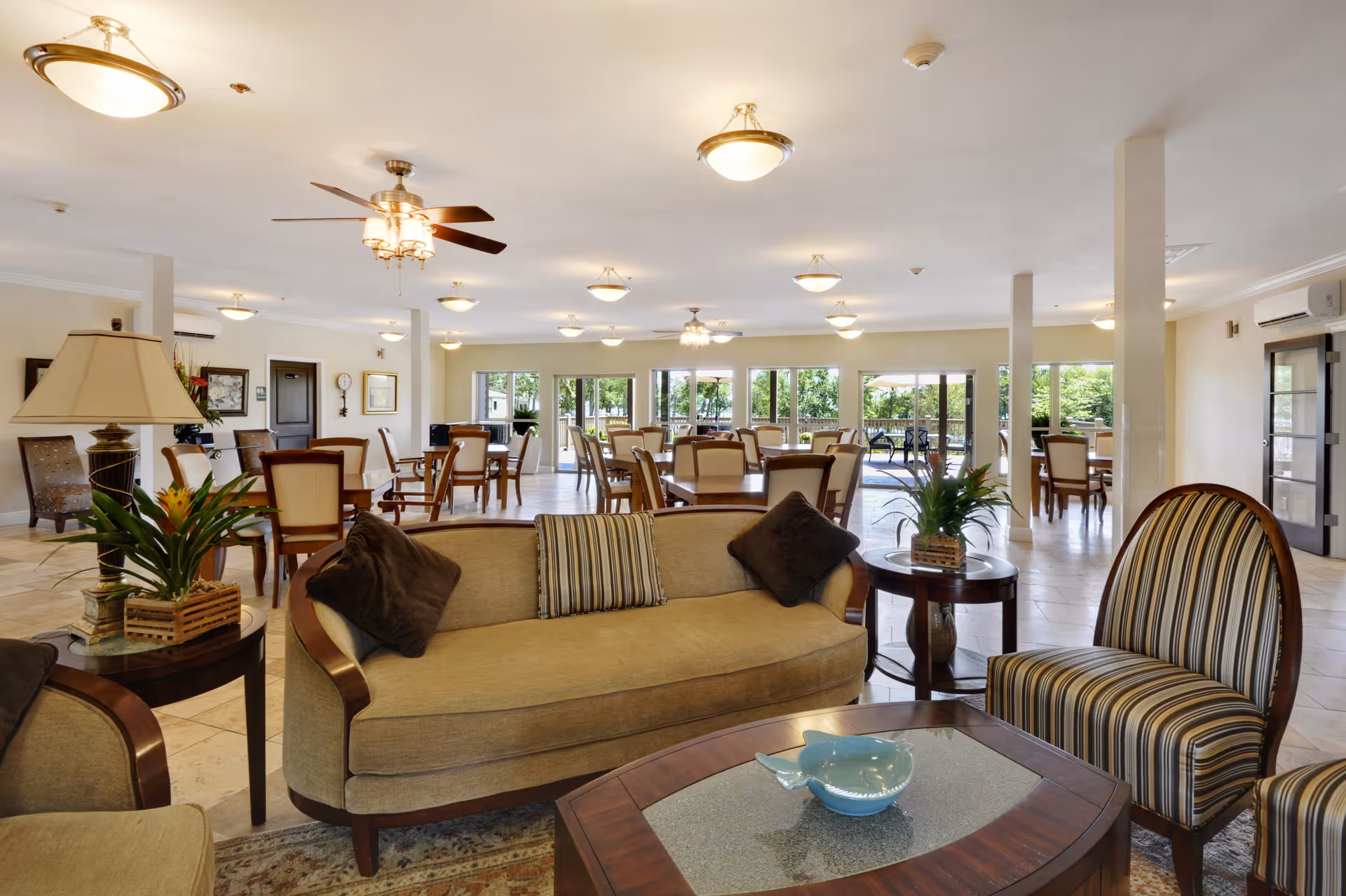 Spacious, well-lit communal living and dining area with sofas, chairs, tables, and large windows overlooking greenery.