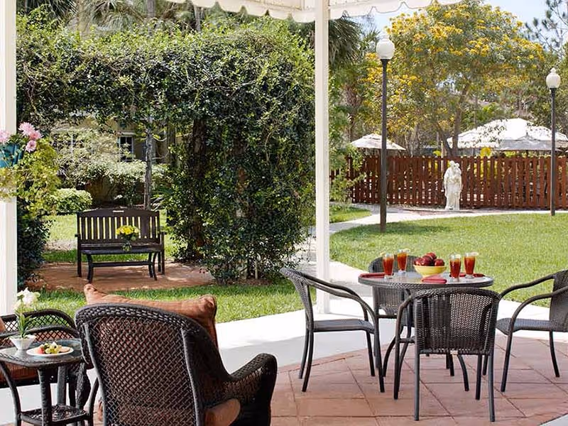 Outdoor patio area with a round table set with glasses of iced tea and a bowl of apples, surrounded by four wicker chairs. In the background, there is a wooden bench with a small flower arrangement, green bushes, trees, and a statue near a wooden fence. The area is shaded by a white canopy.
