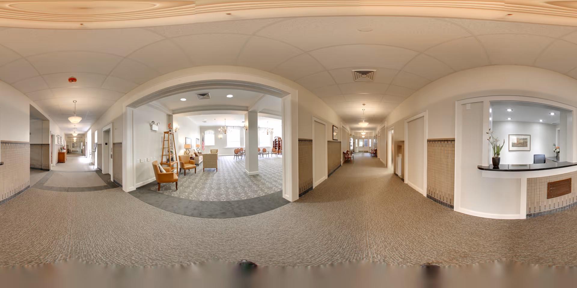 A wide panoramic view of a senior living facility interior hallway with beige carpeting and light-colored walls. On the right side, there is a reception desk with a small plant and framed artwork behind it. In the center, an open area with chairs and tables is visible, illuminated by natural light from large windows. The hallway extends into the distance with doors and light fixtures along the walls.