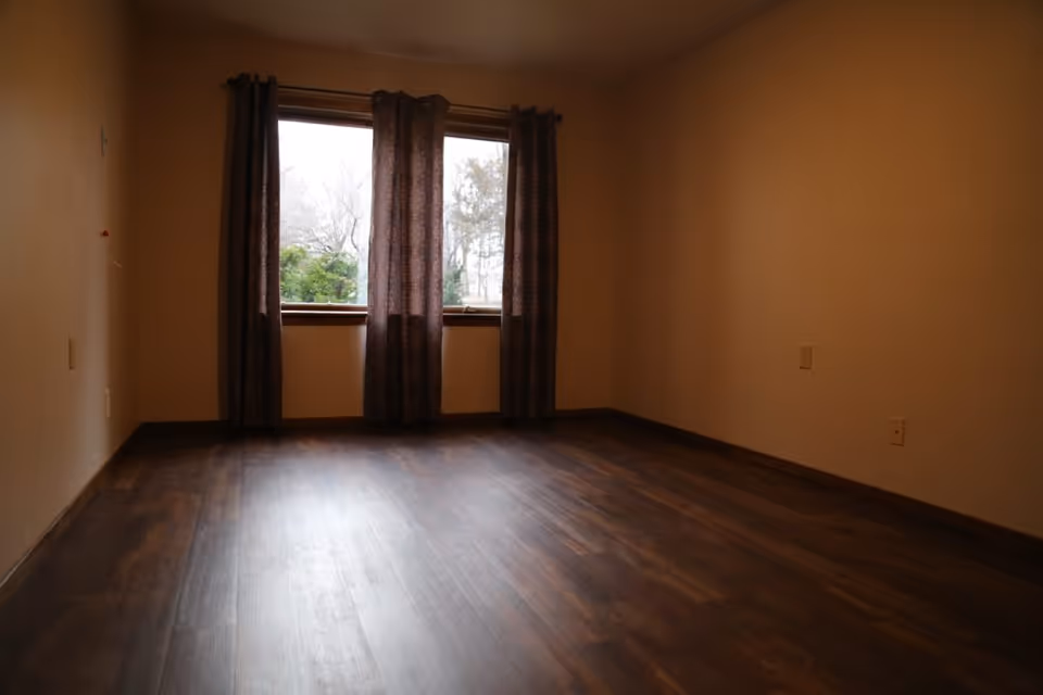 Empty room with dark wooden floor, beige walls, and a window with dark curtains partially drawn, showing some trees outside.