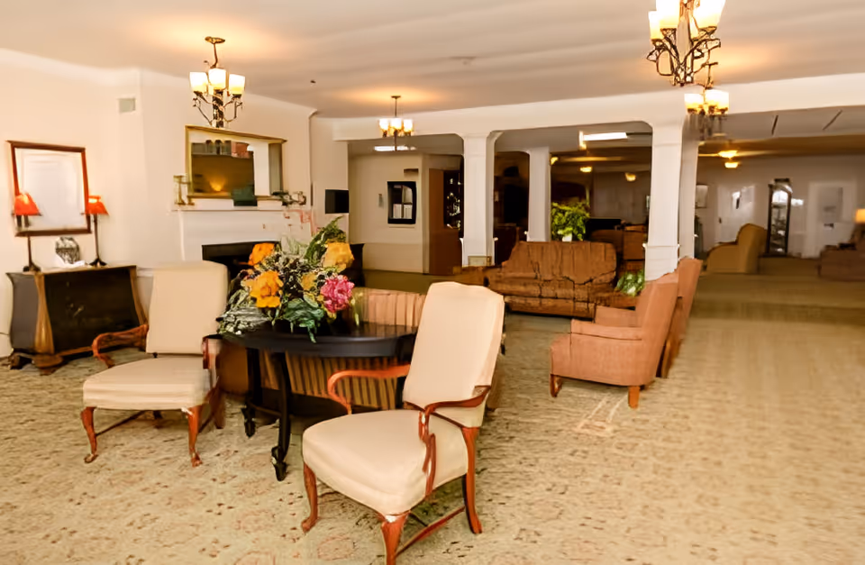 A spacious living room area with multiple upholstered chairs and sofas arranged around a central table adorned with a floral arrangement. The room features a carpeted floor, white walls, a fireplace with a mirror above it, and several ceiling light fixtures providing warm lighting.
