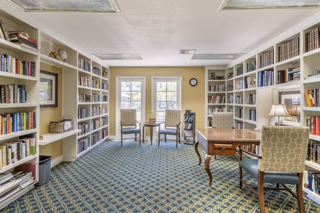 A cozy library room with built-in white bookshelves filled with books lining the walls. There is a patterned carpet on the floor and a wooden table with four upholstered chairs. Two additional chairs and a small table are positioned near two large windows letting in natural light. A clock and decorative items are also visible on the shelves.