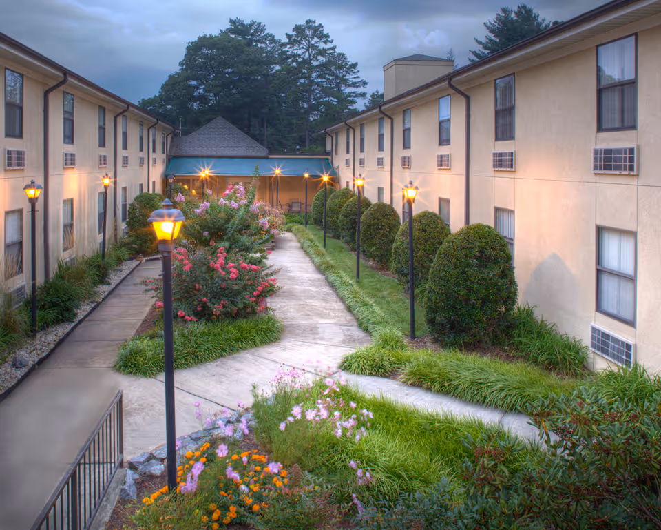 Outdoor courtyard area between two beige multi-story buildings with windows and air conditioning units. The courtyard features a paved walkway, green grass, neatly trimmed bushes, colorful flowers, and several lit lamp posts. Trees and a cloudy sky are visible in the background.