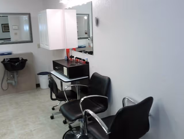 Interior view of a salon area with three black salon chairs with chrome armrests, a black hair washing sink, a white cabinet mounted on the wall, a mirror, and a small table with various hair products on it.