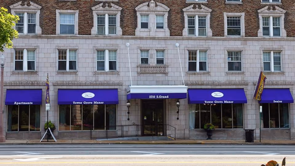 Front exterior view of Tower Grove Manor building with multiple windows and purple awnings above the entrance and windows. The building has a stone facade and is located at 2710 S. Grand.