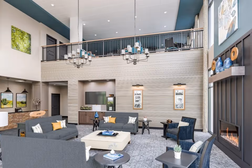 A spacious and modern senior living facility common area with high ceilings, two chandeliers, and a loft railing above. The room features multiple gray sofas and armchairs arranged around a large white ottoman coffee table. There is a fireplace on the right wall with decorative vases on a shelf above it. The walls are decorated with framed pictures and artwork. The floor is carpeted in a light gray pattern, and there is a TV mounted on the far wall above a cabinet.