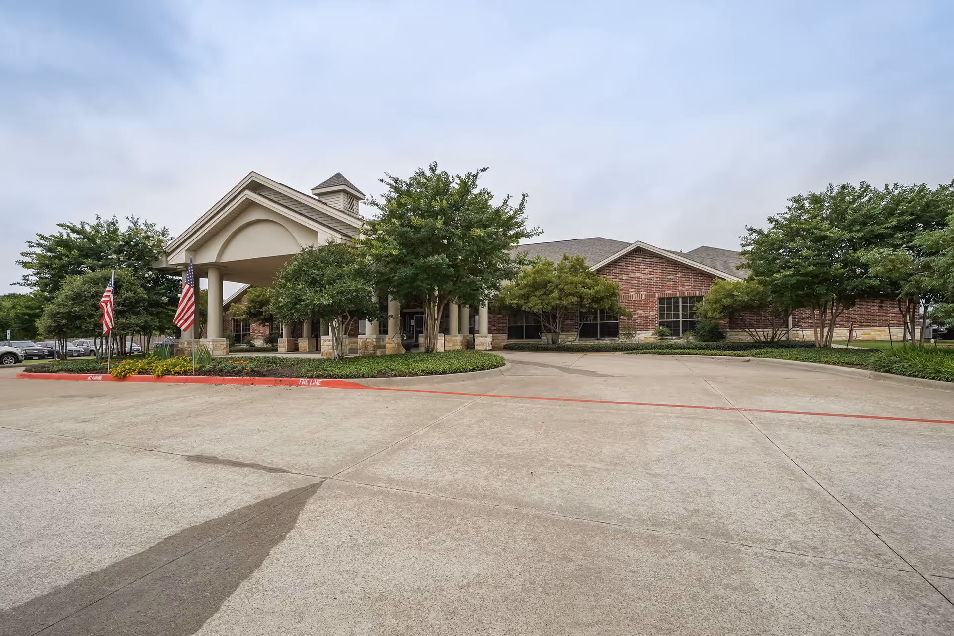 Exterior view of TruCare Living Centers Palestine showing a single-story brick building with a covered entrance supported by columns, surrounded by trees and landscaping, with two American flags displayed near the entrance.