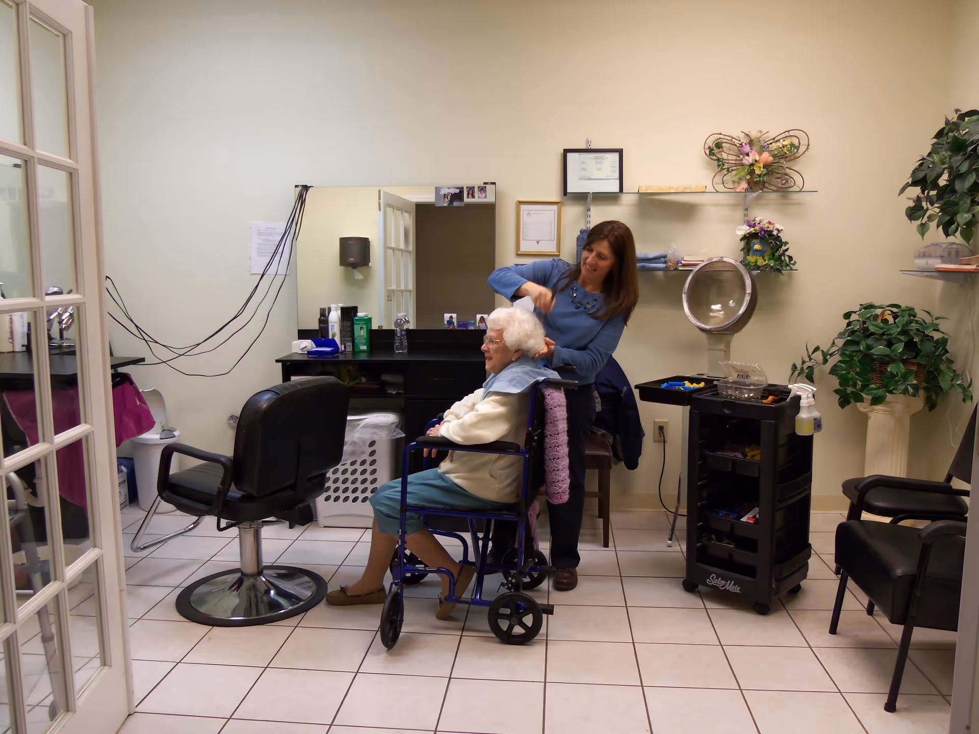 A hair salon room in a senior living facility where a woman is styling the hair of an elderly woman seated in a wheelchair. The room has a large mirror, salon chairs, a hair dryer, shelves with decorative flowers, and a cart with hair styling tools.