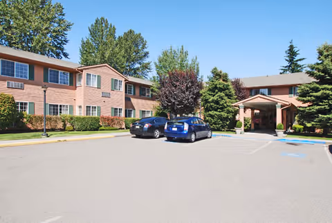 Exterior view of a two-story senior living facility building with a parking lot in front. Two cars are parked near the entrance, which has a covered drop-off area. The building is surrounded by trees and shrubs under a clear blue sky.