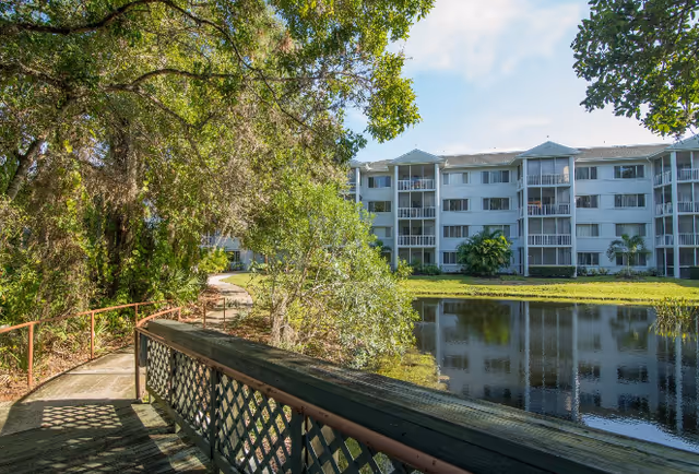 Wooden boardwalk and pathway along a pond with trees and a multi-story residential building in the background.