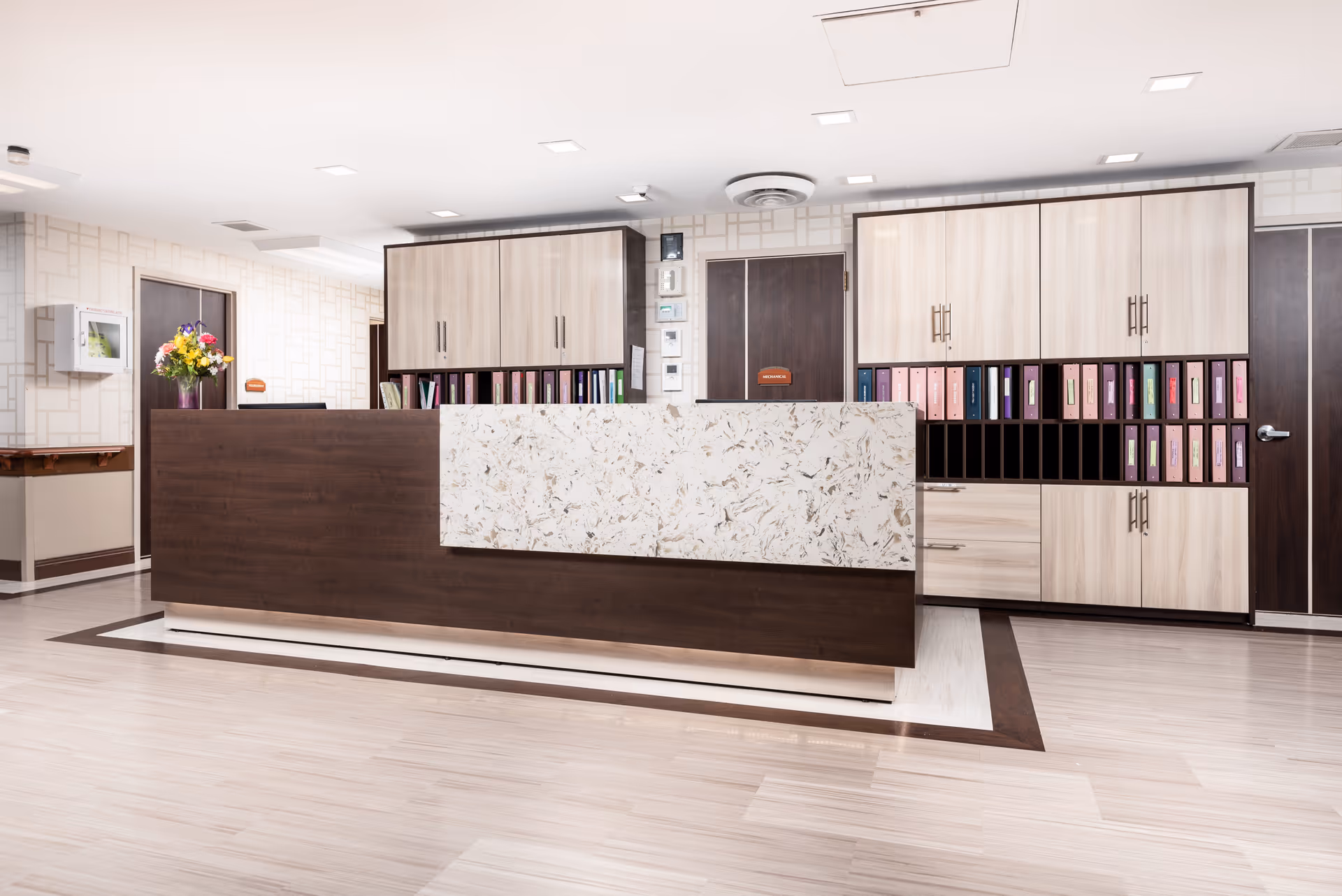 A modern reception desk area in a senior living facility with a dark wood and light marble front counter, light wood cabinets filled with colorful binders, and a vase of flowers on a side table against a patterned wall.