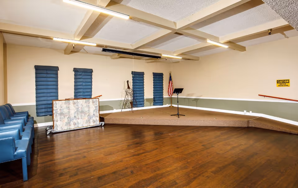 Interior community room with stage, podium, microphone stand, American flag, and blue chairs.