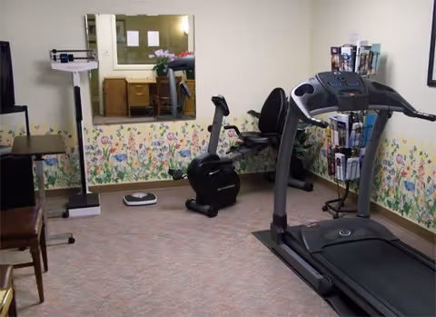 Small fitness room with a treadmill, recumbent exercise bike, scale and a mirror above a floral wallpaper border.