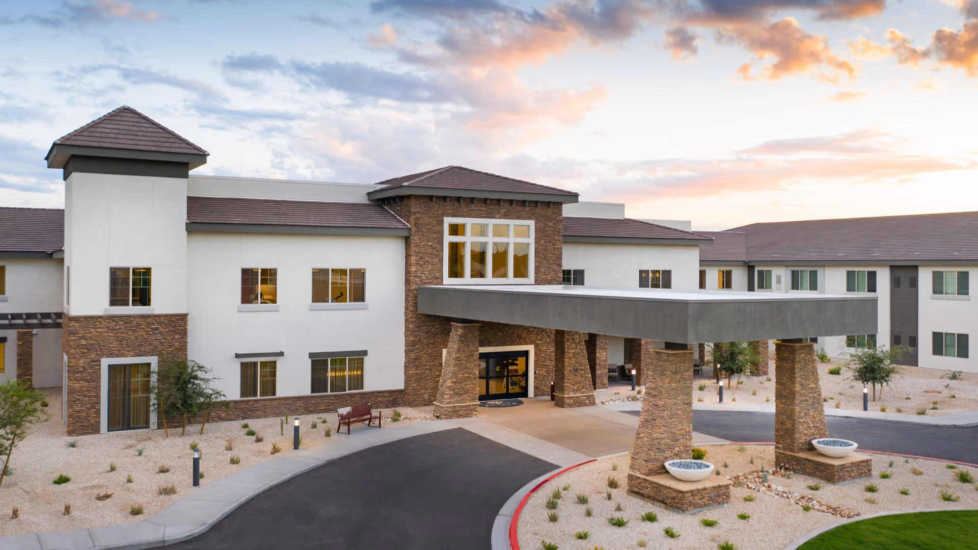 Front exterior of ACOYA Mesa senior living building with a covered porte-cochere, stone columns and desert landscaping at sunset.