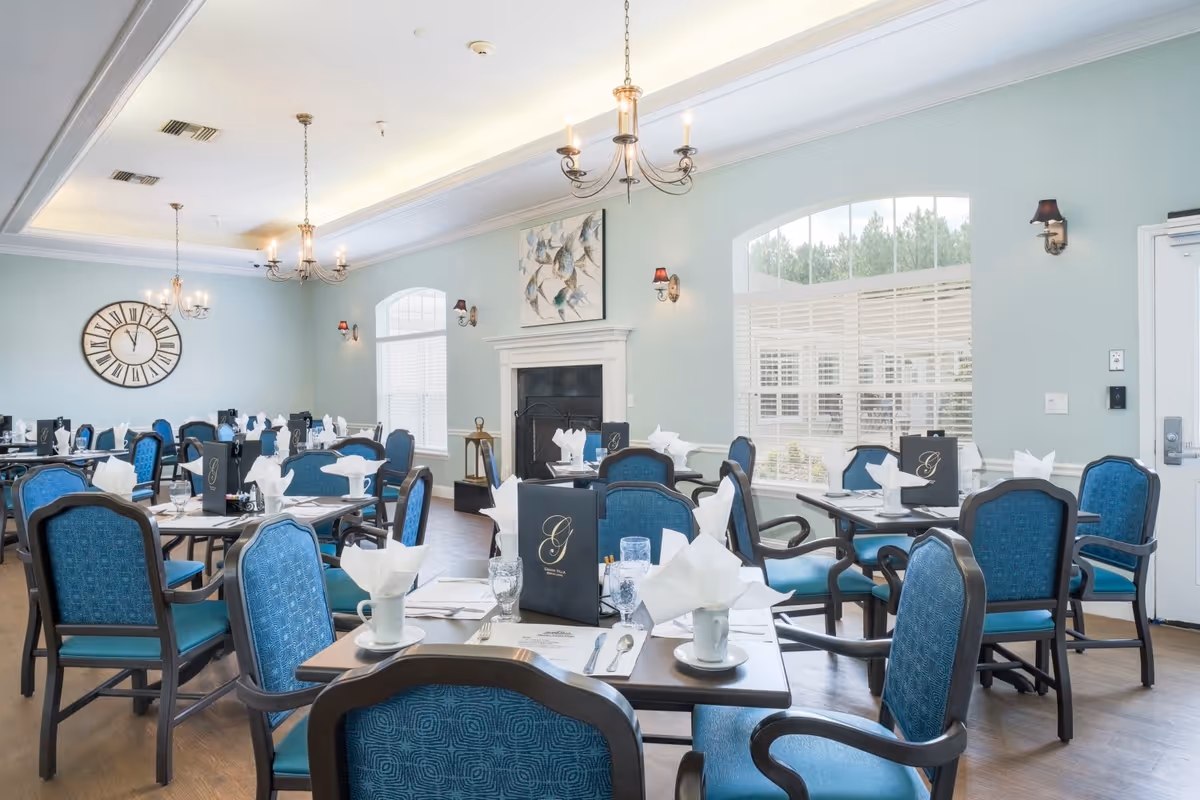 Bright, elegant dining room with multiple set tables and blue upholstered chairs, chandeliers, a fireplace and a large wall clock.