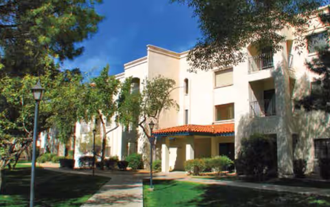 Exterior view of a multi-story residential building with white walls and red-tiled roof accents, surrounded by trees and a well-maintained lawn with a paved walkway and lamp posts under a clear blue sky.