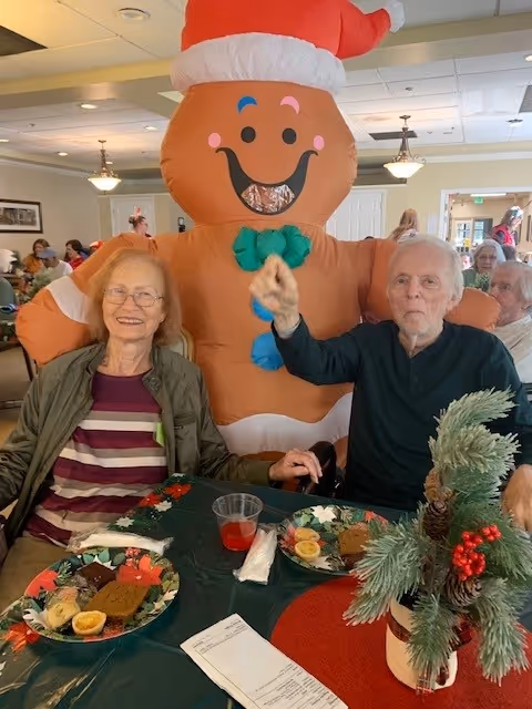 Two elderly individuals sitting at a table with holiday-themed plates of food in front of them. Behind them stands a person in a large inflatable gingerbread man costume wearing a Santa hat. The setting appears to be a festive dining or common area with other people visible in the background.