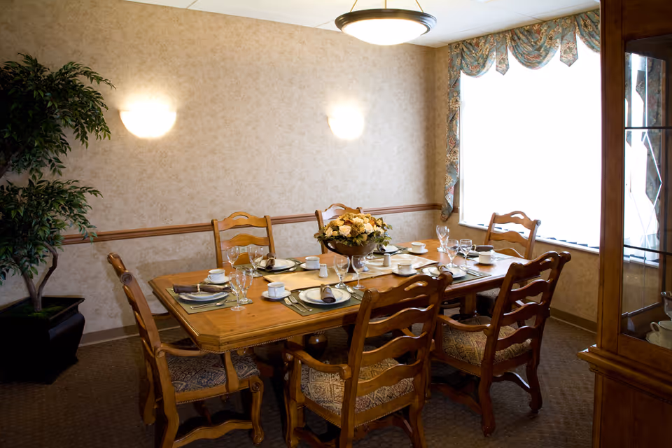 A dining room with a wooden table set for six people, including plates, cups, glasses, and napkins. There is a floral centerpiece on the table. The room has beige wallpaper, a large window with floral curtains, a potted plant in the corner, and a wooden cabinet with glass doors on the right side.