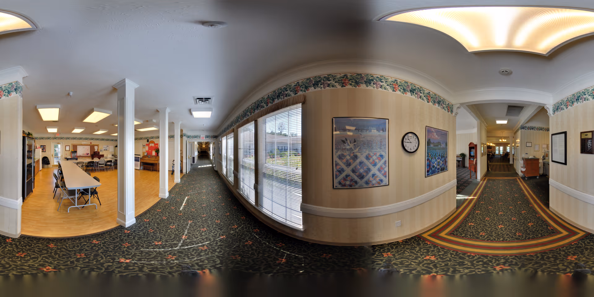 Panoramic view of an interior hallway in a senior living facility with patterned carpet and floral wallpaper border. On the left side, there is a dining area with tables and chairs on a wooden floor. The hallway has large windows with blinds letting in natural light. The walls are decorated with framed pictures and a clock. The ceiling has recessed lighting and a decorative light fixture.