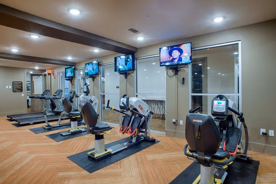Spacious fitness room with several recumbent exercise bikes and treadmills arranged in rows facing wall-mounted televisions.