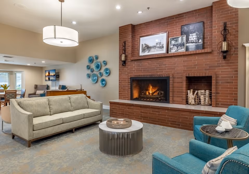 A cozy living room area in a senior living facility featuring a beige sofa, two teal armchairs with striped pillows, a round coffee table, and a brick fireplace with a fire burning. Above the fireplace are three framed black and white photographs and two wall-mounted lanterns. The room has a beige carpet and walls, with a modern ceiling light fixture and decorative blue wall plates on one wall. In the background, there is a dining area with a table, chairs, and a TV mounted on the wall.
