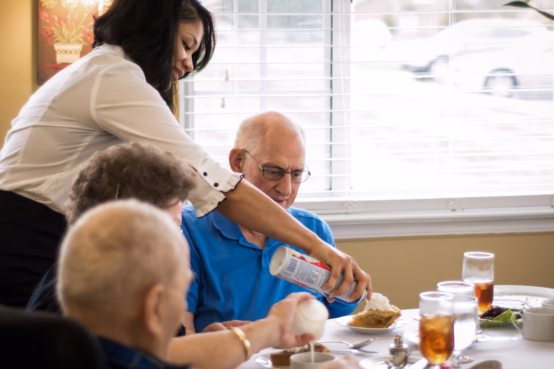 A staff member helps serve whipped cream onto a resident's dessert while several seniors sit at a dining table with drinks.