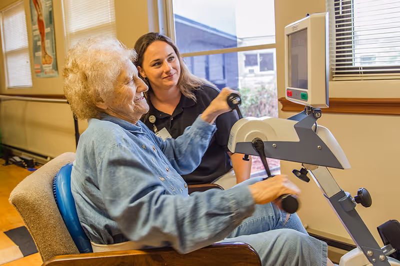 An elderly woman using a hand pedal exercise machine while a caregiver or therapist watches and smiles beside her in a well-lit room with windows and anatomical posters on the wall.