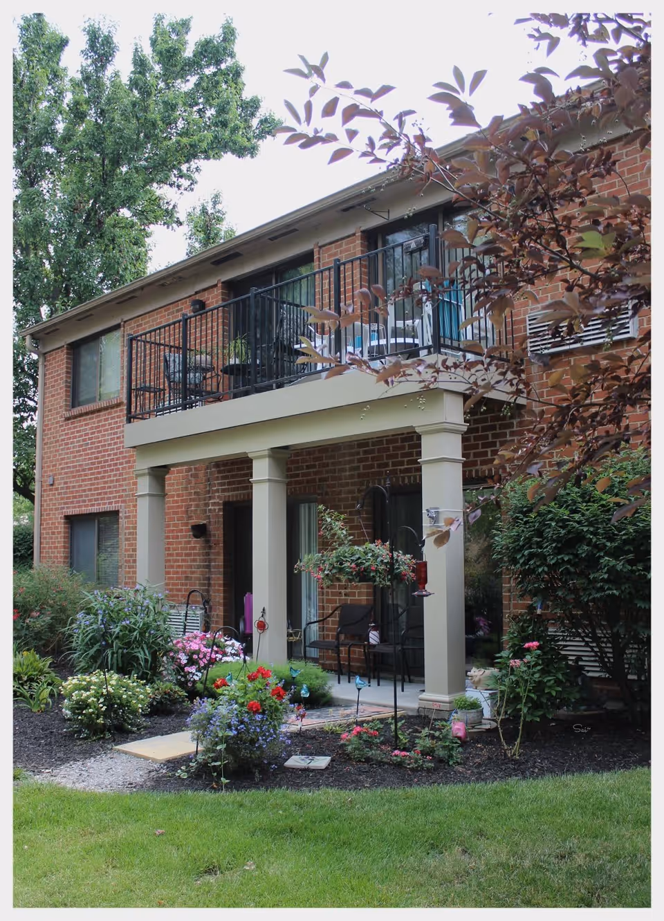 Exterior view of a two-story brick senior living facility with a balcony and a covered patio area. The patio is furnished with chairs and surrounded by a well-maintained garden with various flowering plants and shrubs. Trees and greenery are visible around the building.