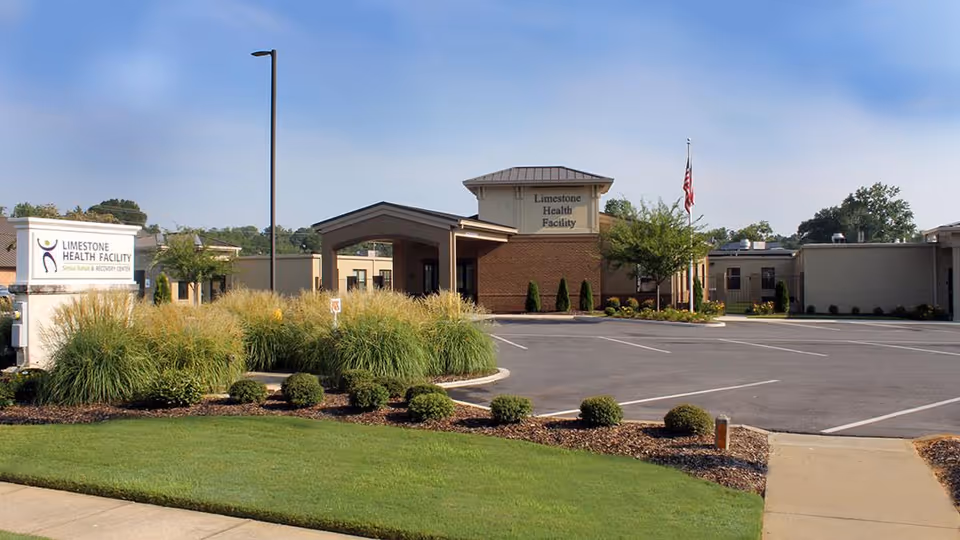 Exterior view of Limestone Health Facility showing the main entrance with a covered drop-off area, a parking lot, landscaped bushes and grass, and a sign that reads Limestone Health Facility Senior Rehab & Recovery Center. An American flag is visible near the entrance under a clear blue sky.