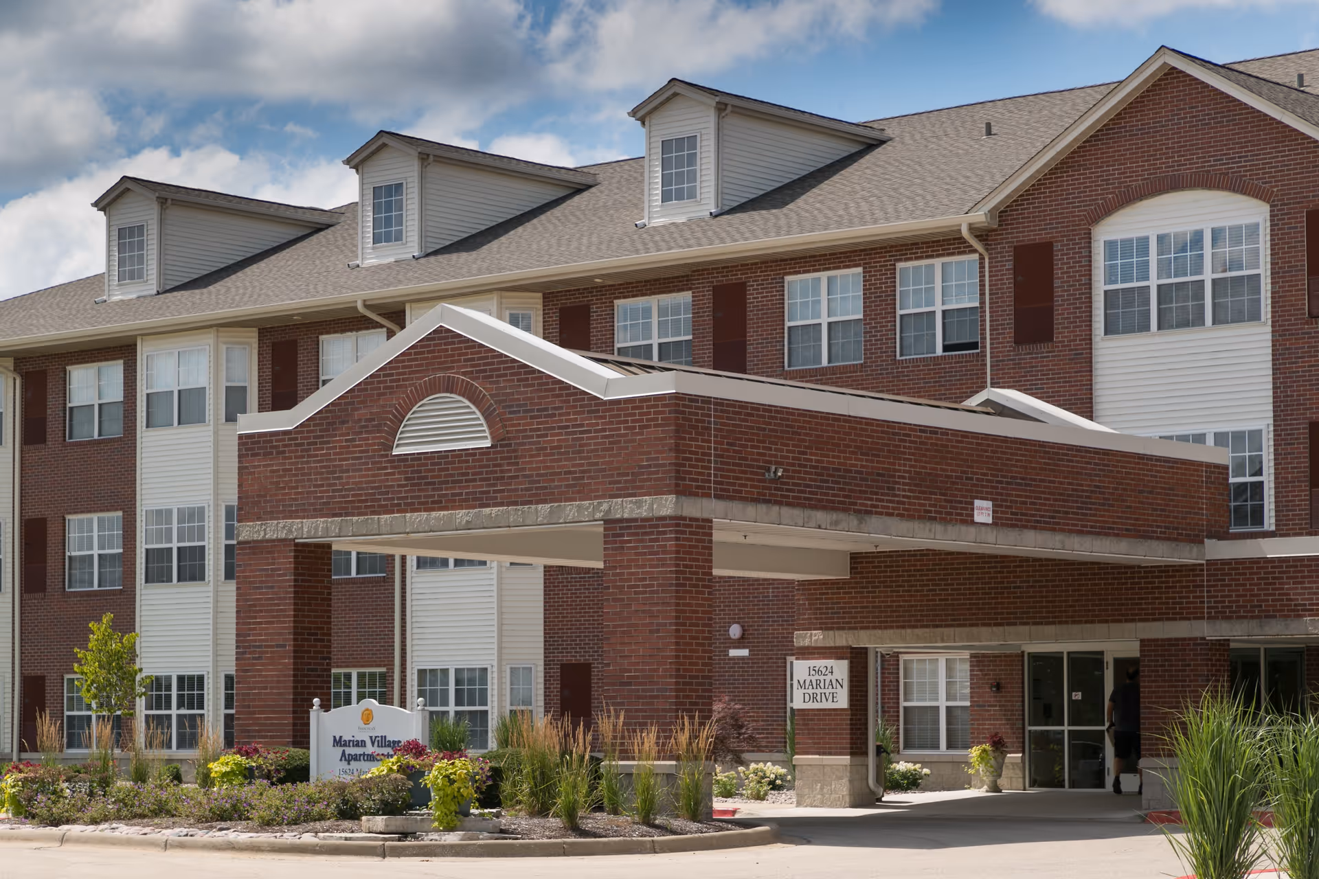 Exterior view of a three-story brick and siding senior living facility building with a covered entrance, landscaped garden, and a sign that reads Marian Village Apartments.