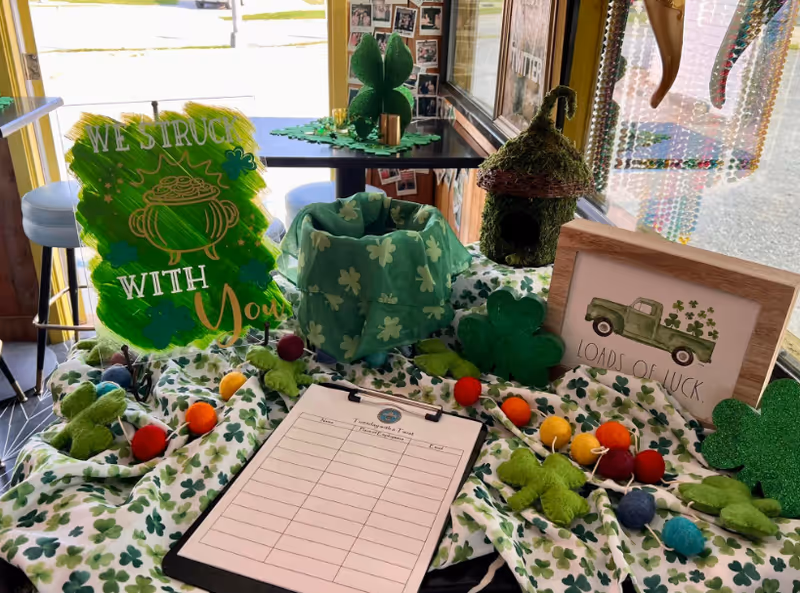 A festive St. Patrick's Day themed table display with shamrock-patterned cloth, green shamrock decorations, colorful felt balls, a moss-covered birdhouse, and a framed picture of a green truck with shamrocks and the text 'Loads of Luck'. A sign reads 'We struck with you'. A clipboard with a sign-up sheet is also on the table. The display is set near a window with bead curtains and a view outside.