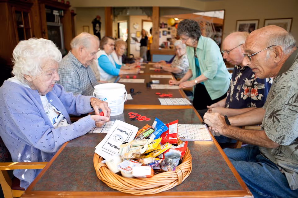 A group of elderly people sitting around a long table playing bingo. The table has bingo cards, red bingo chips, and a basket filled with various snacks and small items. The setting appears to be a communal activity room in an assisted living facility.