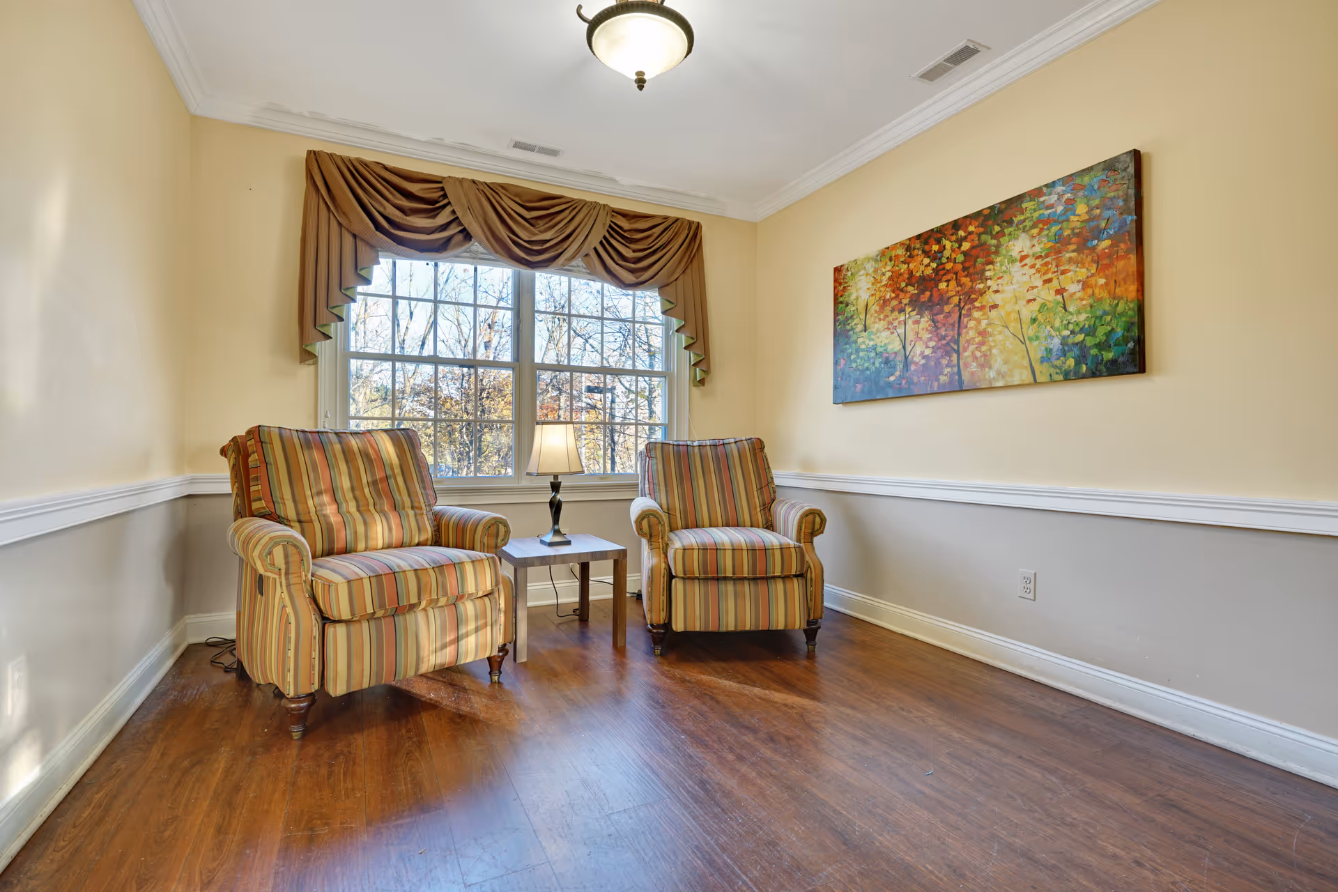 A cozy sitting area with two striped armchairs positioned around a small wooden side table with a lamp. The room has a large window with brown drapes, light yellow walls with white trim, and a colorful abstract painting on the wall. The floor is wooden.