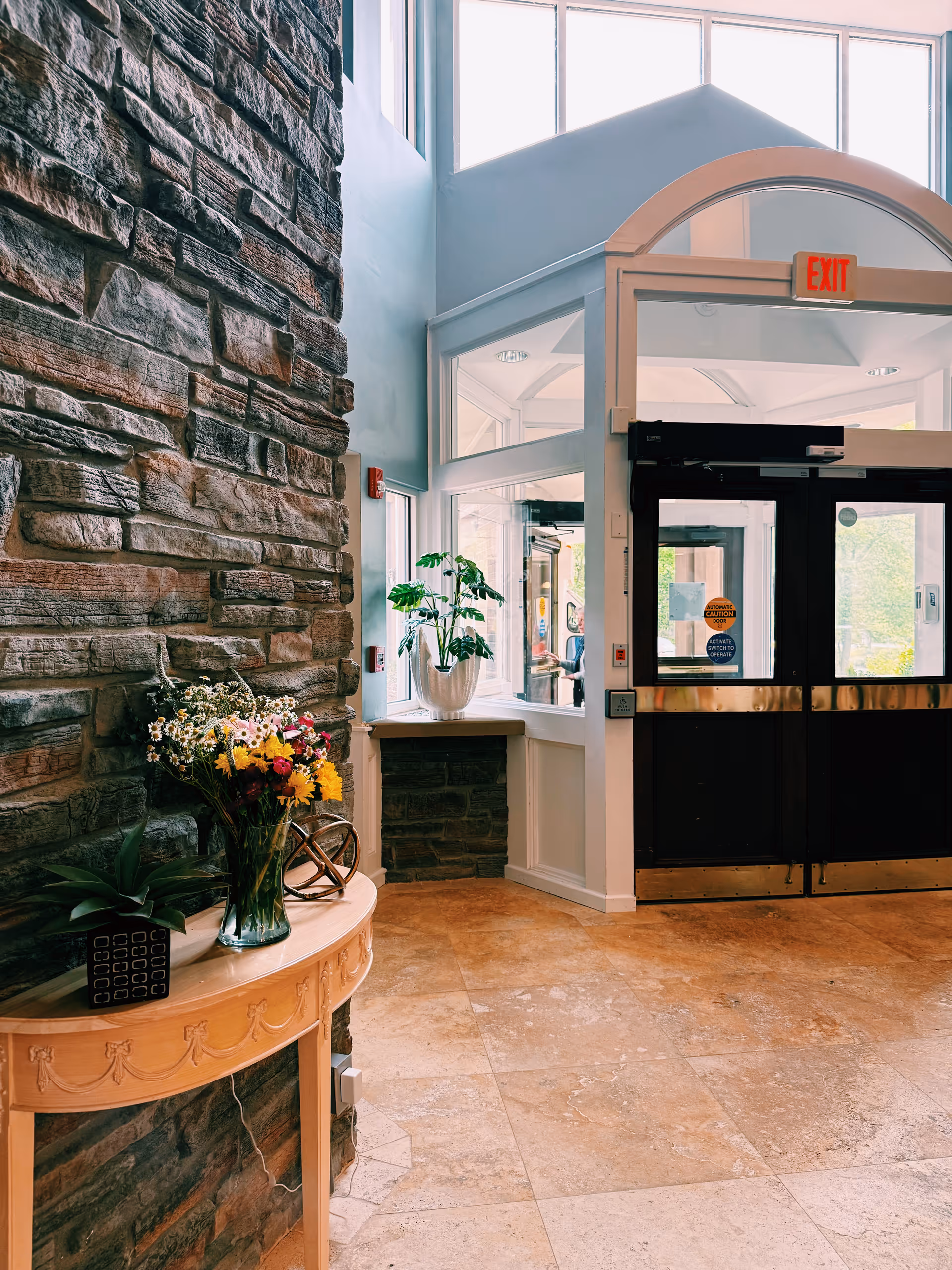 Sunlit entry lobby with a stone accent wall, a console table holding flowers and plants, and double glass exit doors.