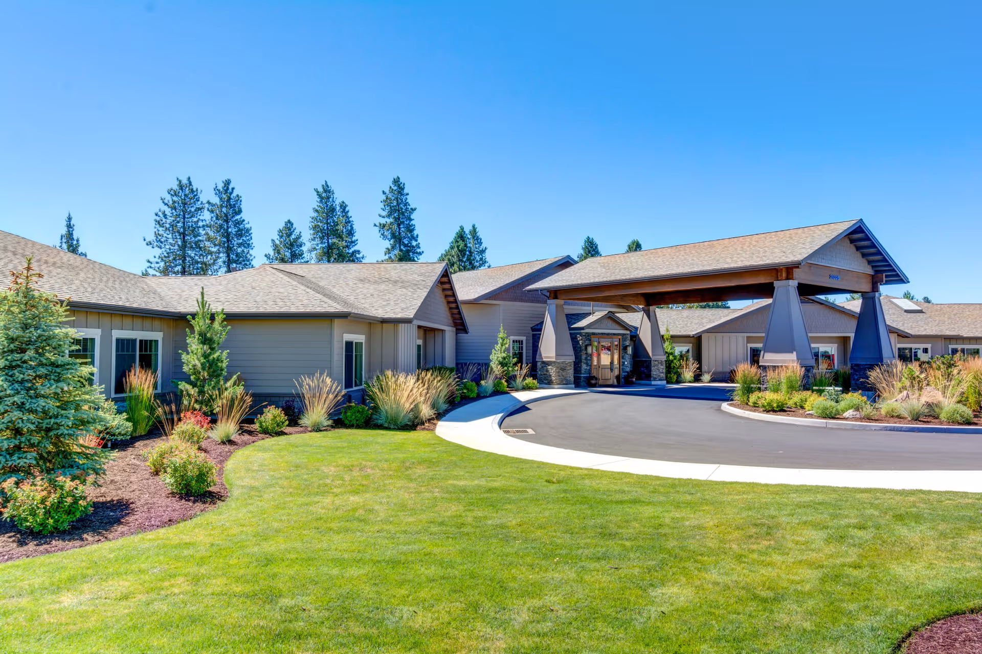 Front entrance and porte-cochère of a single-story assisted living facility with landscaped lawns, shrubs, and pine trees under a clear blue sky.