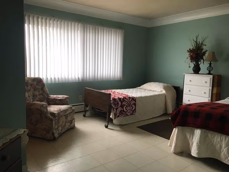 A simple bedroom in a retirement facility with two single beds, one with a red and black checkered blanket and the other with a white quilt and a red patterned throw. There is a floral upholstered armchair near a large window with vertical blinds, and a white dresser with a lamp and a vase of flowers on top against a teal wall.