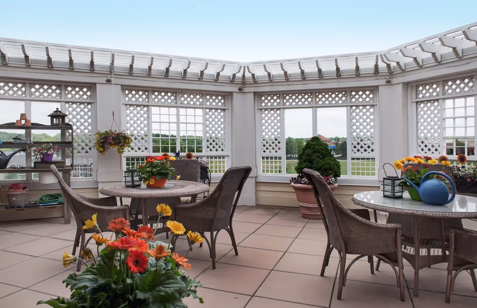A bright and airy enclosed patio area with large windows and white lattice panels. The space features two round tables with wicker chairs around them, decorated with colorful potted flowers and a blue watering can. There are additional plants and gardening supplies on a shelf to the left, and a large potted shrub near the windows. The floor is tiled, and the ceiling has a white pergola-style structure.