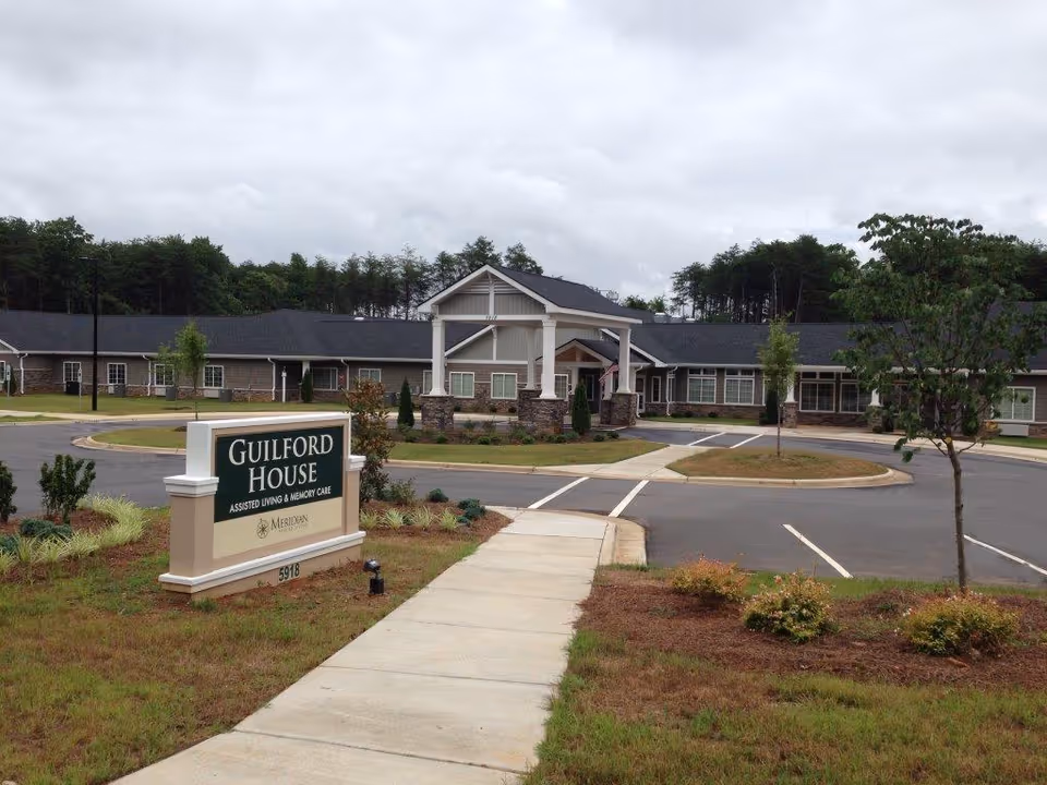 Exterior view of Guilford House assisted living and memory care facility showing a single-story building with a covered entrance, surrounded by a parking lot, landscaped grass, small trees, and shrubs under a cloudy sky.