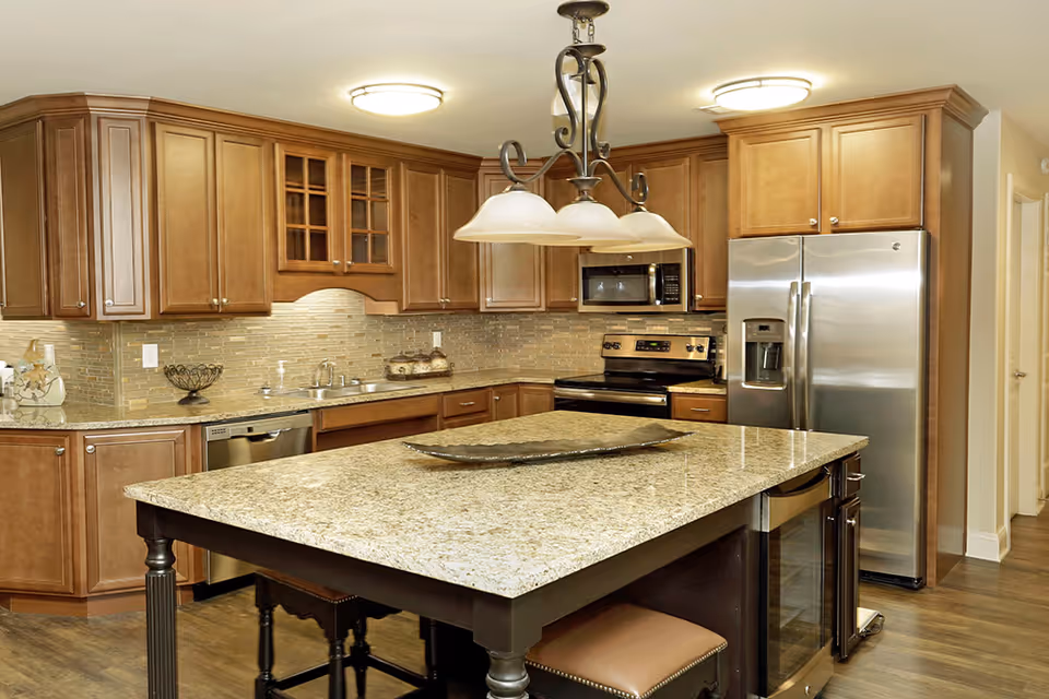 A modern kitchen with wooden cabinets, a large granite island with seating, stainless steel appliances including a refrigerator, oven, microwave, and dishwasher, and a decorative light fixture hanging above the island.