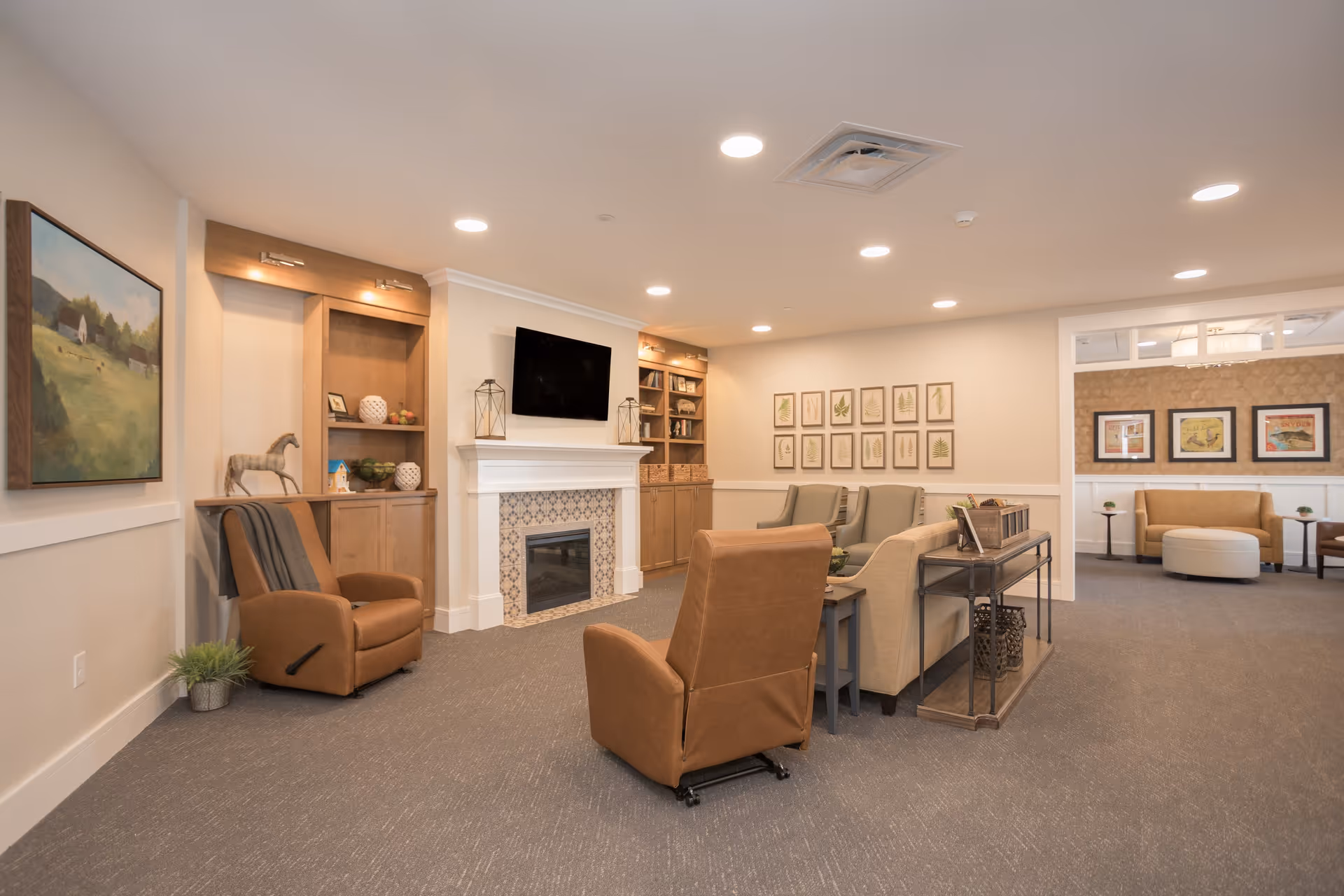 A cozy living room area in a senior living facility with beige walls, carpeted floor, and recessed lighting. The room features a fireplace with a mounted flat-screen TV above it, built-in wooden shelves with decorative items, and comfortable seating including brown leather recliners and beige armchairs. There are framed botanical prints on one wall and a painting of a countryside scene on another. An adjacent room with additional seating and framed artwork is visible through an open doorway.