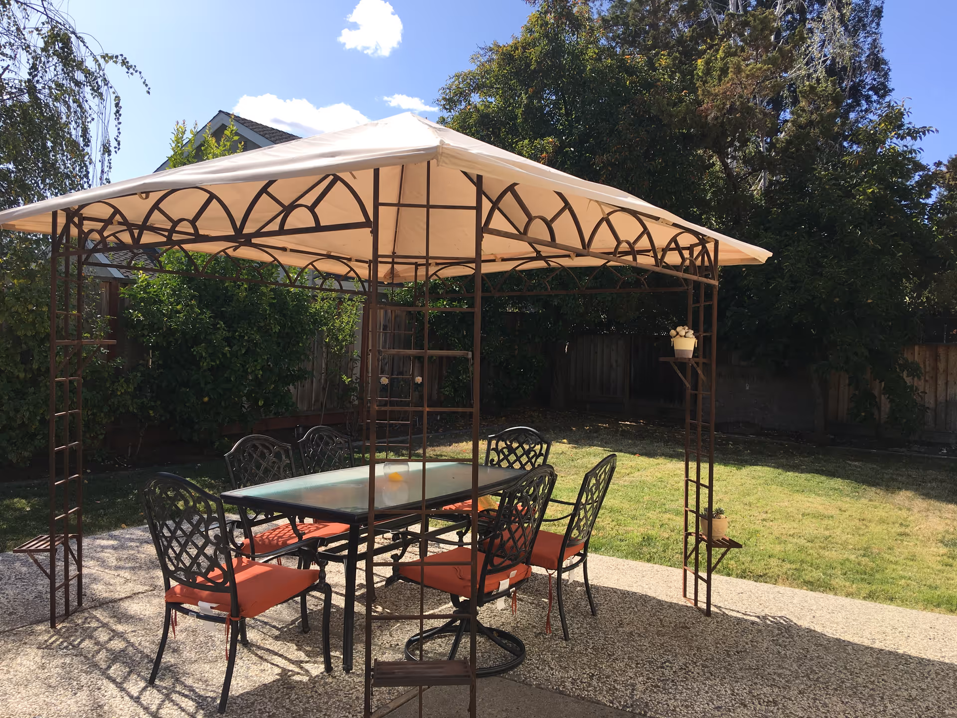 Outdoor patio area with a metal-framed gazebo covered by a beige canopy. Underneath the gazebo is a glass-top table surrounded by six metal chairs with red cushions. The patio is set on a gravel surface with a grassy yard and trees in the background under a clear blue sky.
